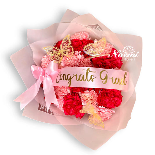 24 claveles en tonos de rosa y rojo, con delicados detalles de mariposas, cinta personalizada y un lazo decorativo - Floristería Noemi Flowers.